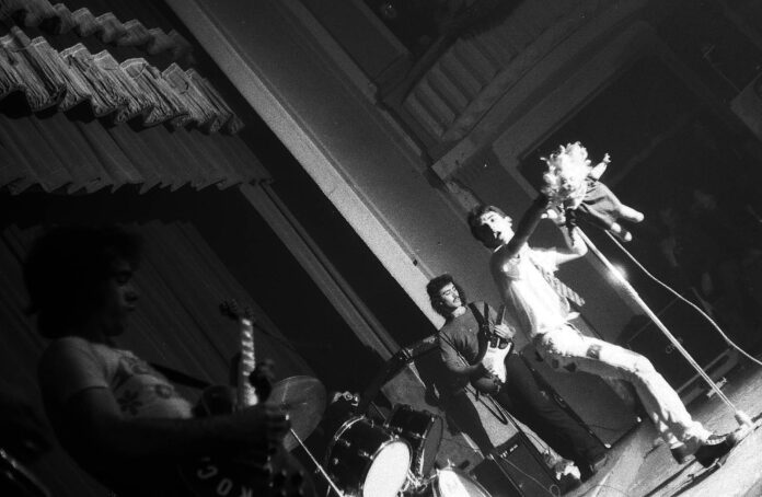 Fotografia en blanc i negre de Ramoncín actuant amb els seus músics sobre l'escenari del Festival Punk de l'Aliança del Poblenou, 4 de desembre de 1977. Foto de Mercè Salvador.